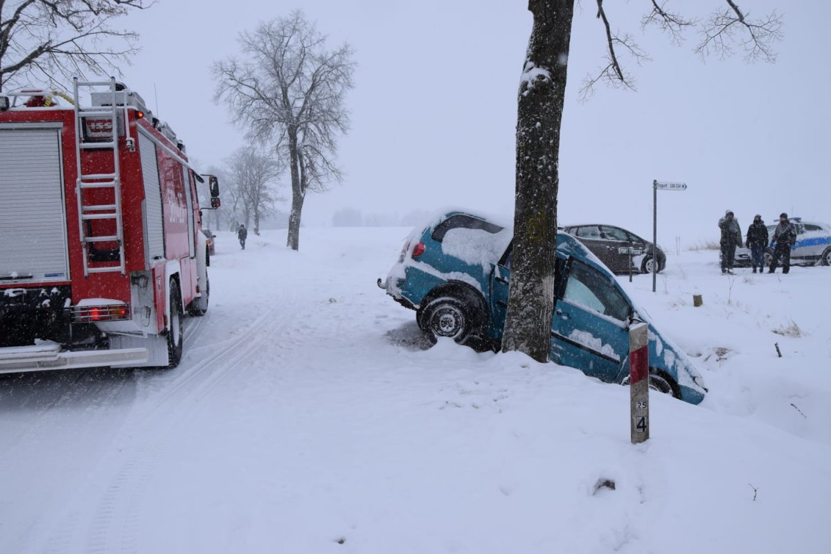 Po lewej stronie na zaśnieżonej drodze stoi wóz strażacki, po prawej stronie w rowie stoi samochód, dalej radiowóz przy którym stoją trzy osoby i samochód