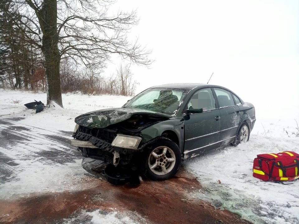 Uszkodzony pojazd stojący bokiem na drodze po lewej stronie stoi drzewo po prawej na ziemi leży torba medyczna