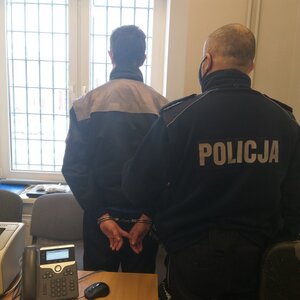 Dwóch mężczyzn stojących w pomieszczeniu tyłem. Jeden to umundurowany policjant, drugi zatrzymany mężczyzna, który ma założone kajdanki na ręce trzymane z tyłu