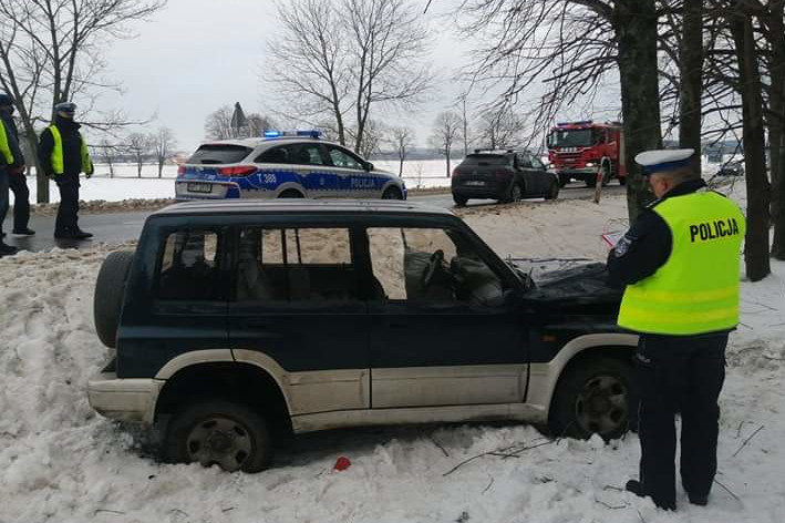 Na zdjęciu na poboczu widać pojazd który uderzył w drzewo, na drodze stoją służby ratownicze, przy samochodzie stoją policjanci
