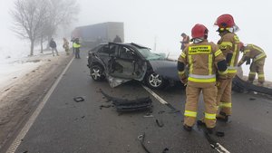 na środku drogi rozbity samochód osobowy przy nim służby ratownicze, w tle samochód ciężarowy