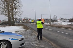 Policjant stojący przy drodze z urządzeniem do pomiaru prędkości, w tle budynki