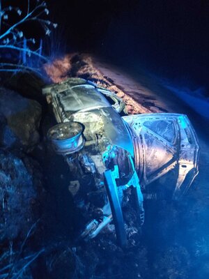 Spalony samochód z otwartą pokrywą silnika leżący w w rowie na boku