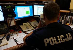 Oficer dyżurny siedzi przy biurku, przed monitorami i rozmawia przez telefon,
