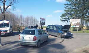 2 pojazdy rozbite na skrzyżowaniu dróg, w tle służby ratownicze