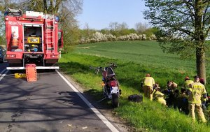 Po prawej stronie w rowie strażacy udzielają pomocy osobie, po lewej stronie na drodze wóz strażacki i motocykl