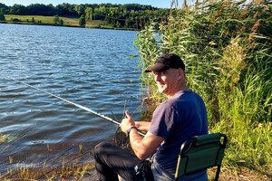 Pomysłodawca zawodów na stanowisku wędkarskim. Siedzi na krześle przy jeziorze z wędką w dłoniach