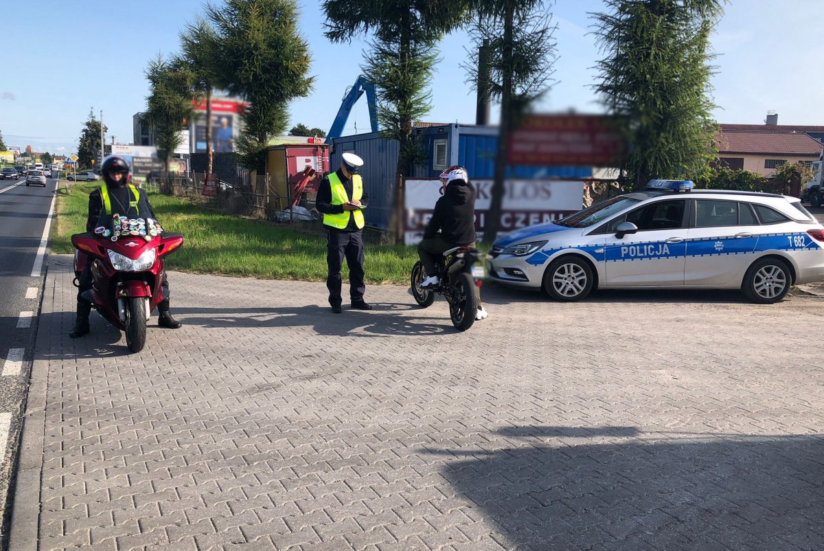 Policjant podczas kontroli drogowej dwóch motocyklistów. Dwa motocykle i motocykliści oraz radiowóz na placu obok drogi