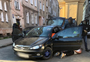 Moment zatrzymania mężczyzny podejrzanego o kradzież rozbójniczą. Zatrzymany mężczyzna leży na ziemi obok swojego samochodu, a nad nim stoją policjanci.