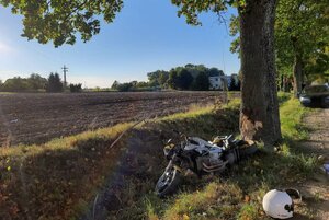 Miejsce zdarzenia drogowego. Rozbity motocykl leżący przy drzewie i samochód stojący na drodze (na drugim planie)