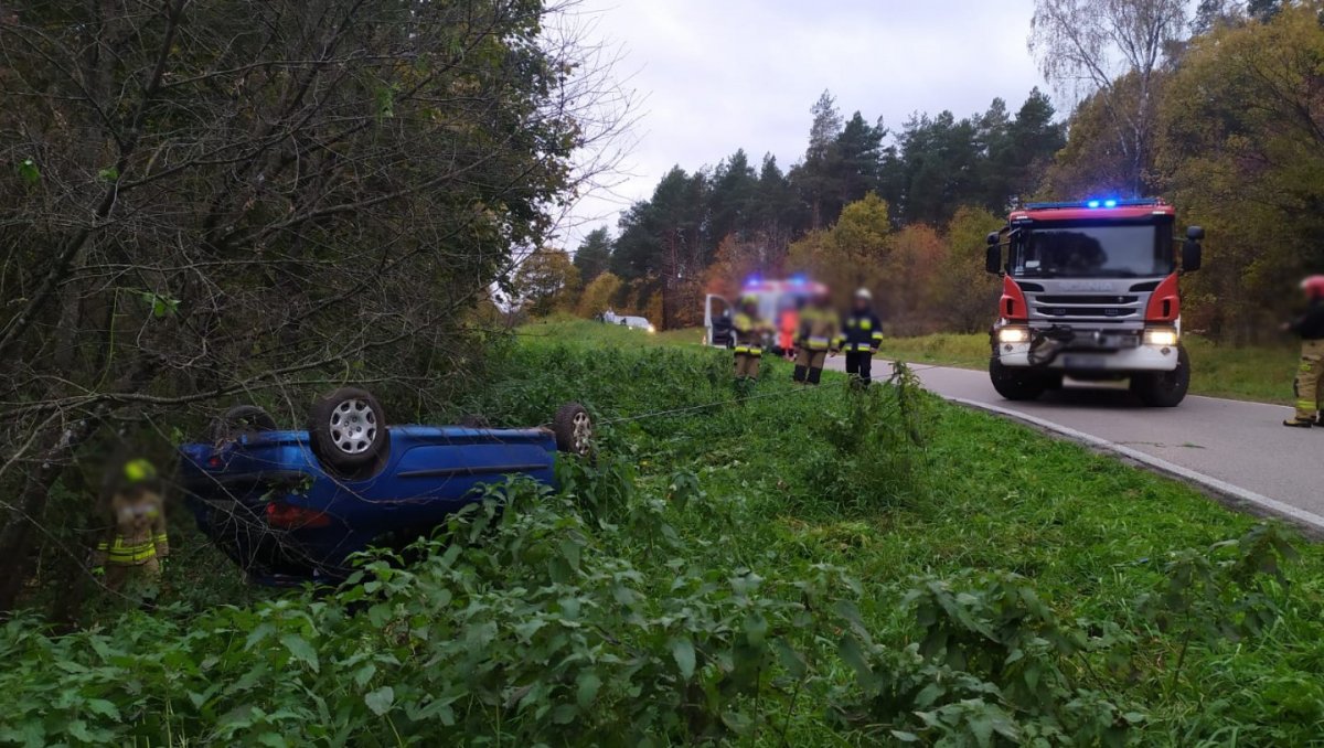 Rozbity samochód leżący w rowie i wóz strażacki oraz strażacy na drodze