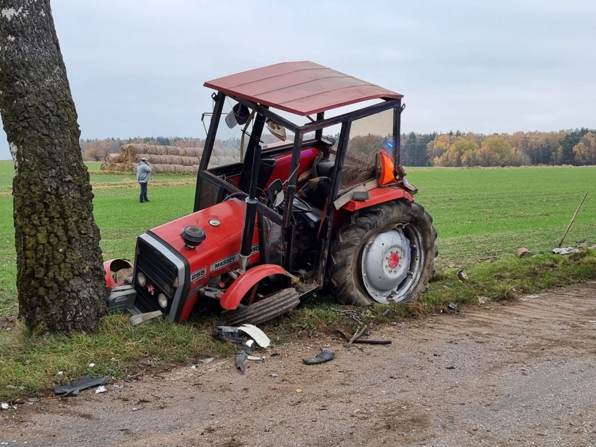 uszkodzony ciągnik rolniczy koloru czerwonego stojący na poboczu przed drzewem, po uprzednim uderzeniu w drzewo