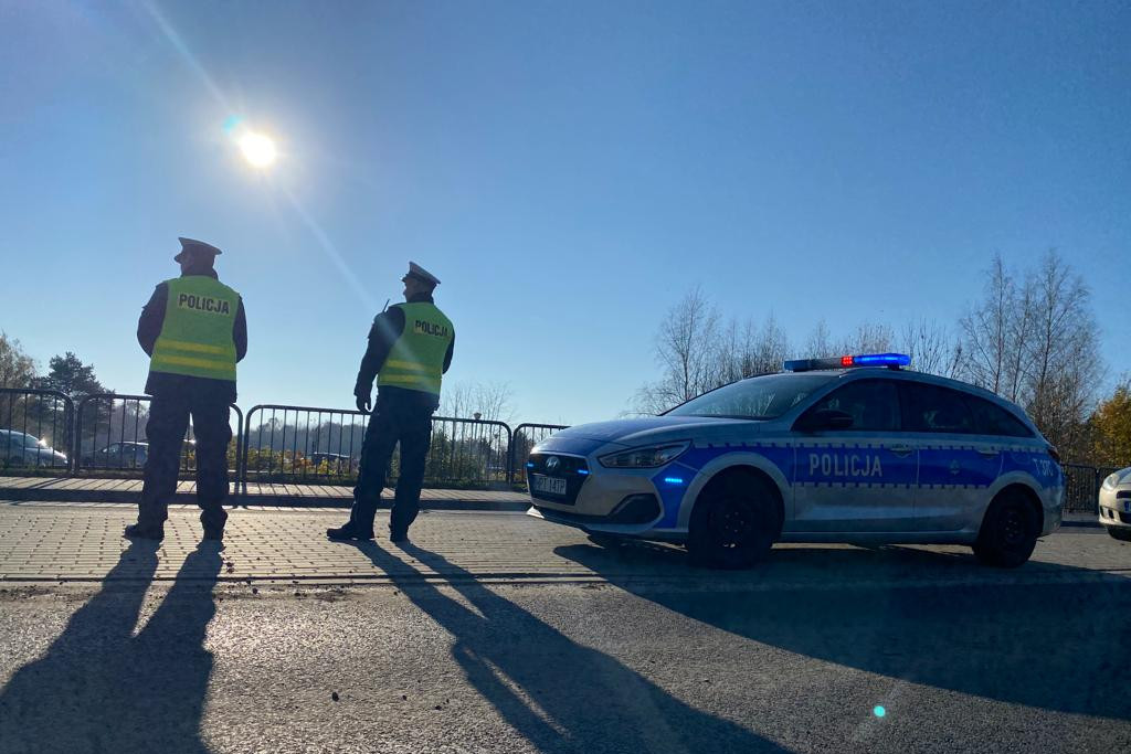 Dwoje policjantów stojących na drodze obok radiowozu na tle niebieskiego nieba i świecącego słońca