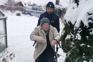 Policjant przemieszczając się przez zaśnieżoną dróżkę pomaga przemieścić się mężczyźnie siedzącemu w wózku ułatwiającym mu poruszanie się.