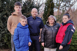 Policjant z synem oraz rodziną, którą przyjął pod swój dach stojący na trawniku na tle drzew