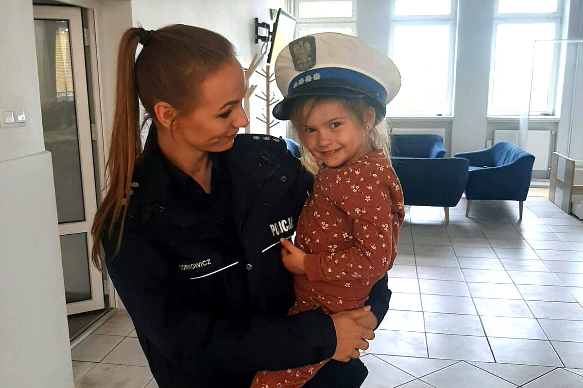 Policjantka trzyma na rękach małą dziewczynkę, która ma na głowę założoną policyjną czapkę.