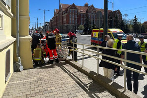Miejsce zdarzenia drogowego. Policjanci i ratownicy wykonujący czynności wobec poszkodowanych na chodniku obok budynku