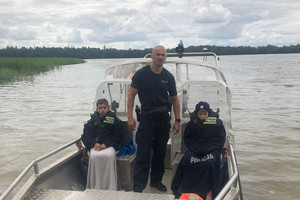 Policjant stoi na pokładzie łodzi, a po jego obu stronach siedzą młodzi chłopcy przykryci policyjnymi mundurami.