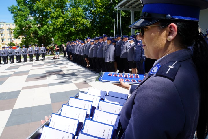 Policjantka na tacy trzyma legitymacje dla odznaczonych a w tle stoją policjanci