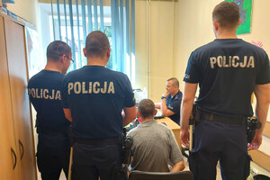 Trzech stojących policjantów, a pomiędzy nimi siedzący zatrzymany. W tle siedzący policjant za biurkiem