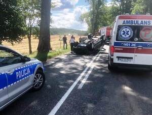 samochód do góry kołami przy nim stojące osoby i karetka pogotowia