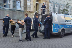 Zatrzymane osoby prowadzone przez policjantów z radiowozu do budynku komendy
