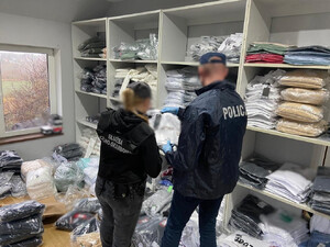 Funkcjonariusze przeszukują pomieszczenie, w którym na podłodze i na regałach poukładane są ubrania w workach foliowych.