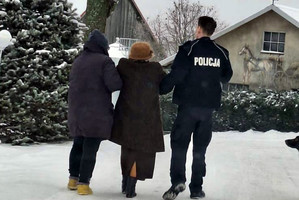 Zgłaszająca i policjant prowadzą seniorkę po zaśnieżonej drodze w kierunku budynków
