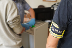 Zdjęcie przedstawia policjantów i zatrzymanego mężczyzny podczas pobierania odcisków palców.