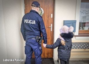 Umundurowany policjant trzyma za rękę dziewczynkę. Obydwoje stoją przed drzwiami