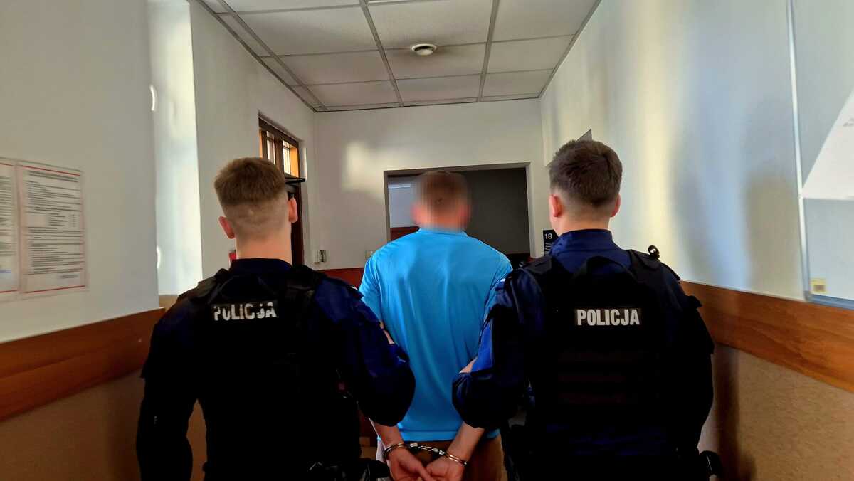 Na zdjęciu widać dwóch policjantów podczas doprowadzenia osoby zatrzymanej. Zdjęcie wykonano w jednostce policji