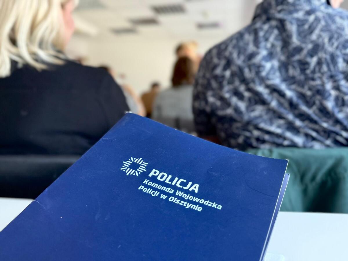 Na pierwszym planie granatowa teczka z napisem Komenda Wojewódzka Policji w Olsztynie. W tle uczestnicy szkolenia.