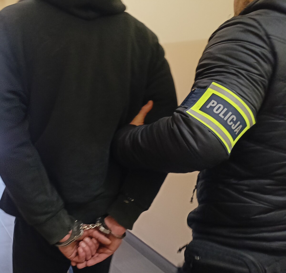 Policjant służby kryminalnej przytrzymuje zatrzymanego, który ma założone kajdanki na ręce trzymane z tyłu