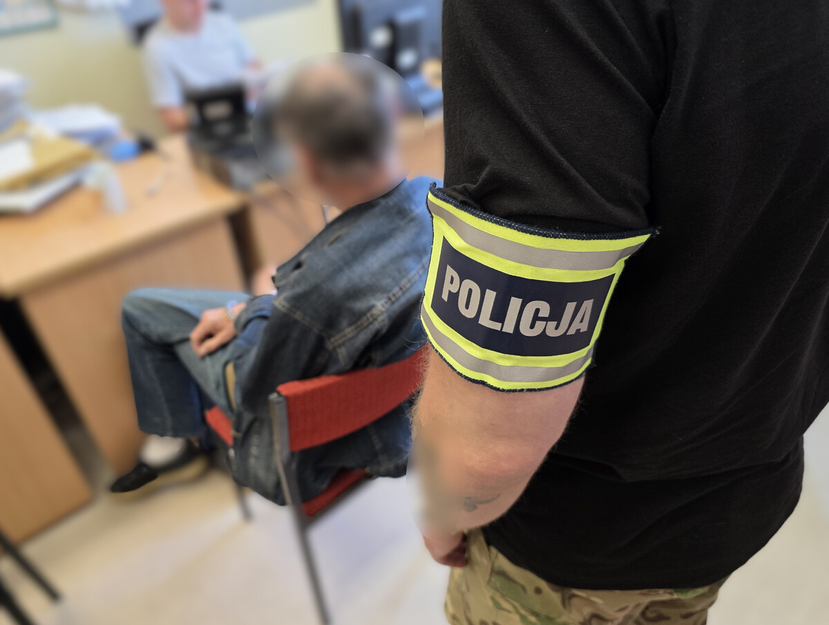 Policjant pionu kryminalnego stoi przy zatrzymanym mężczyźnie. Zatrzymany siedzi na krześle